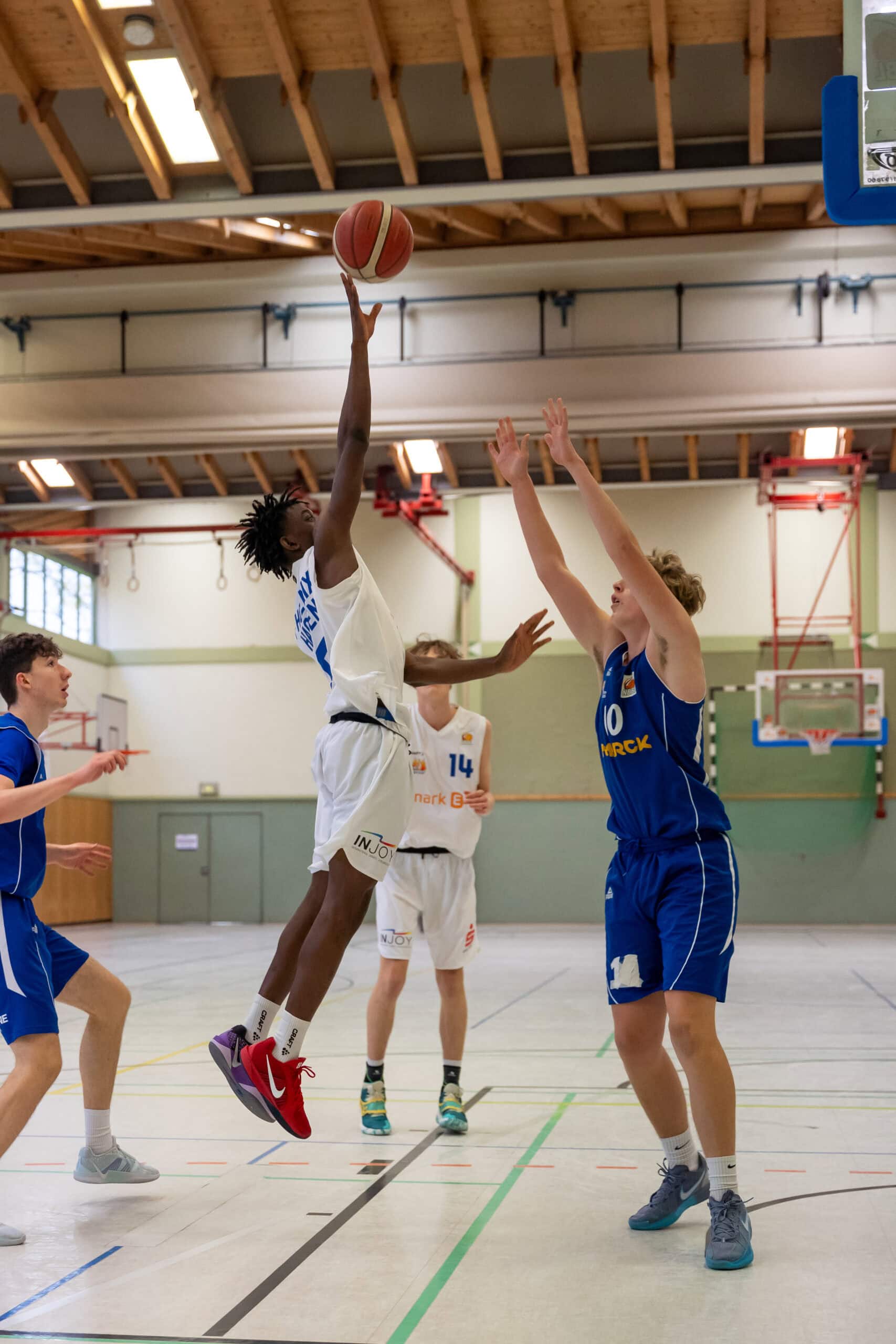 Ein Basketballspieler in Weiß springt, um den Ball zu werfen; andere in blauen Trikots verteidigen.