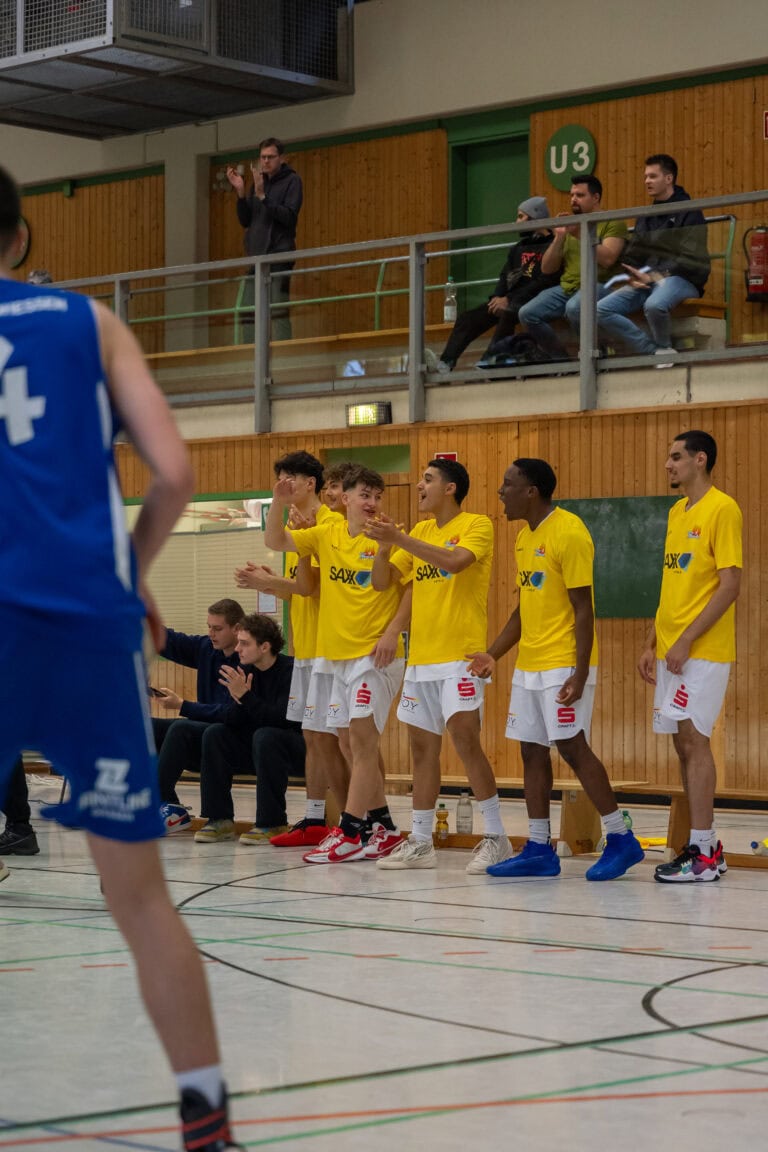 Basketballteam Mitglieder in gelben Trikots jubeln am Spielfeldrand, Spieler in Blau im Vordergrund.