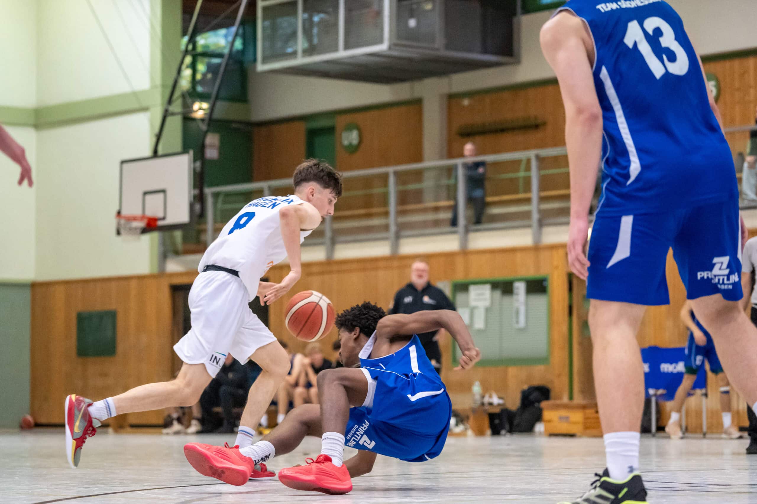 Basketballspieler in Weiß dribbelt Ball, während blauer Spieler rutscht; dynamische Szene in Turnhalle.