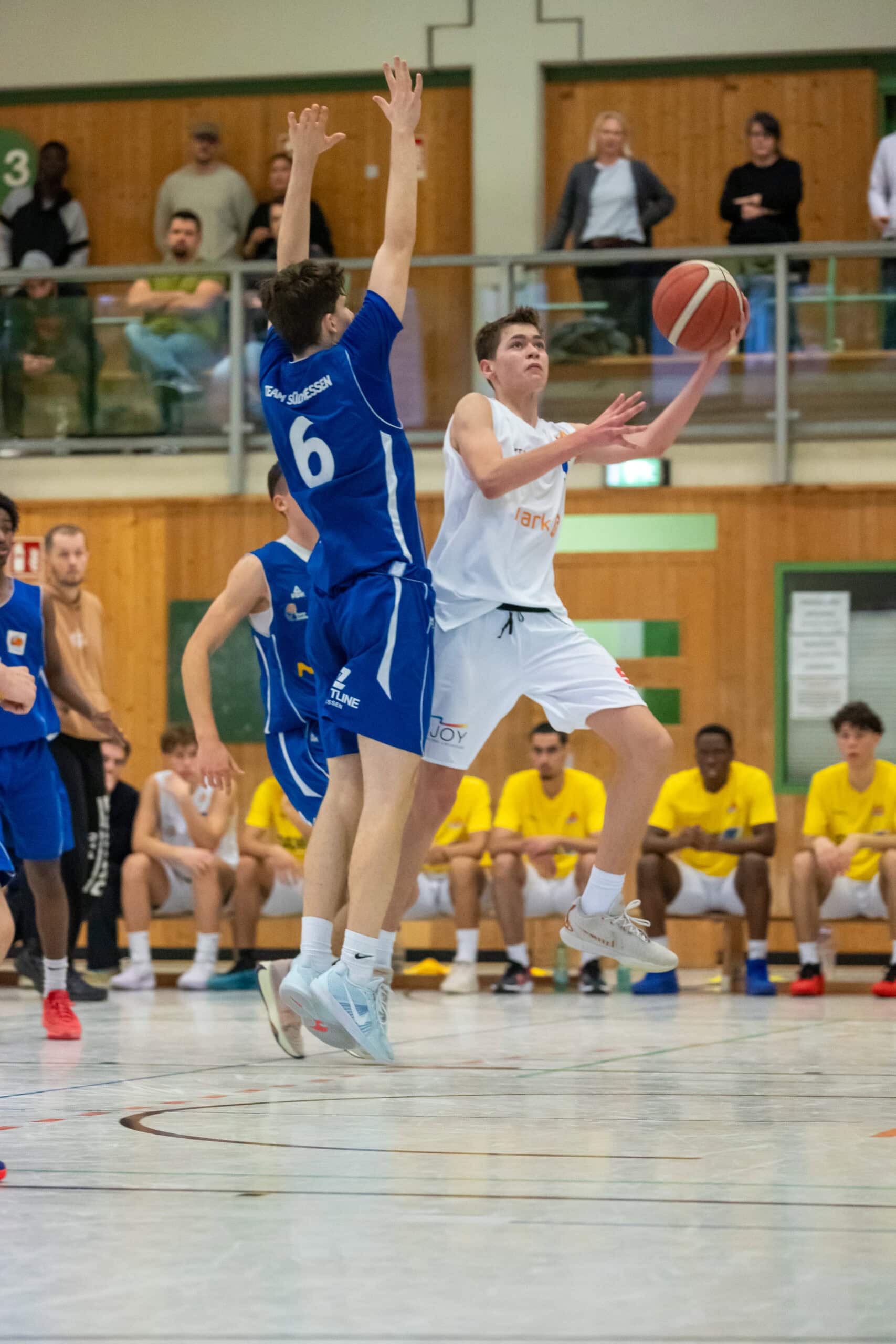 Ein Basketballspieler in weiß springt hoch, um zu werfen, während ein Verteidiger in Blau blockt.