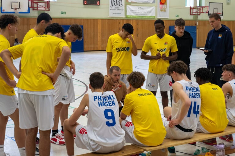 Trainer spricht mit Basketballteam, Spieler tragen gelbe und weiße Trikots, konzentrierte Atmosphäre.