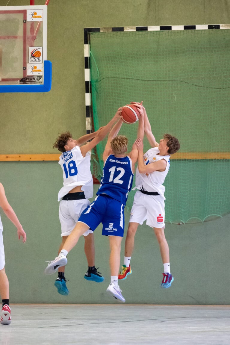 Drei Basketballspieler im Sprung kämpfen um den Ball, konzentrierte Gesichter, blau-weiße Trikots.