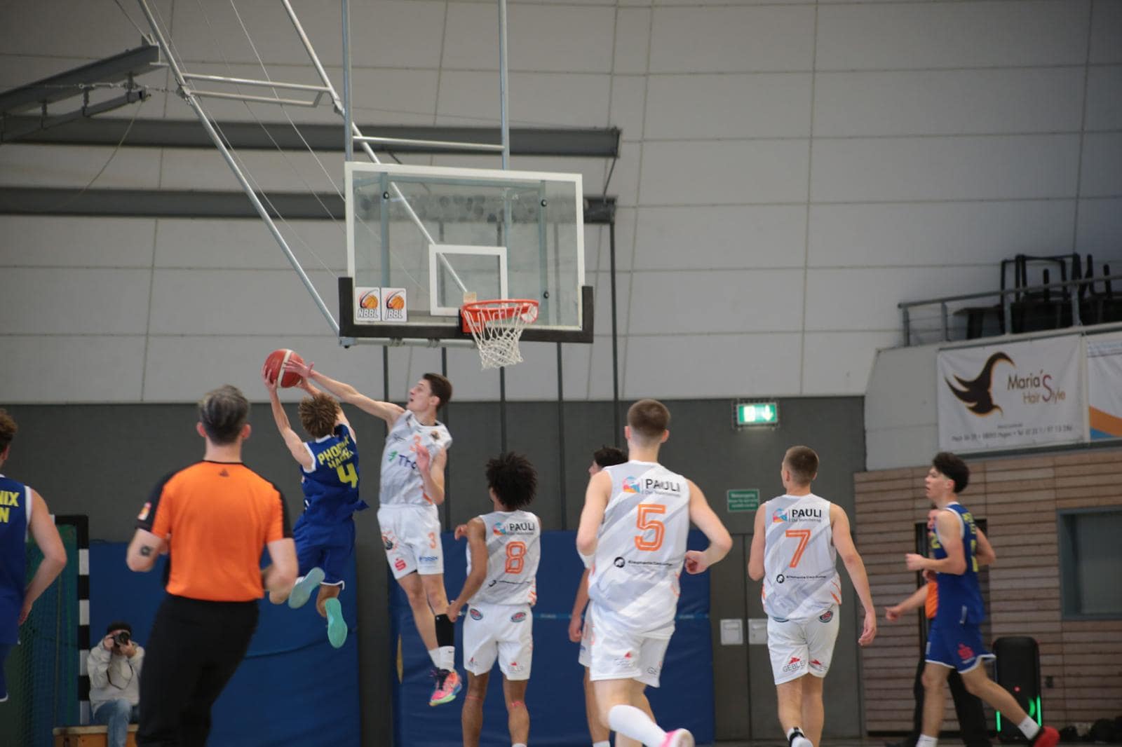 Ein Basketballspiel mit Spielern in weißen Trikots und einem Spieler in blauem Trikot beim Korbwurf.