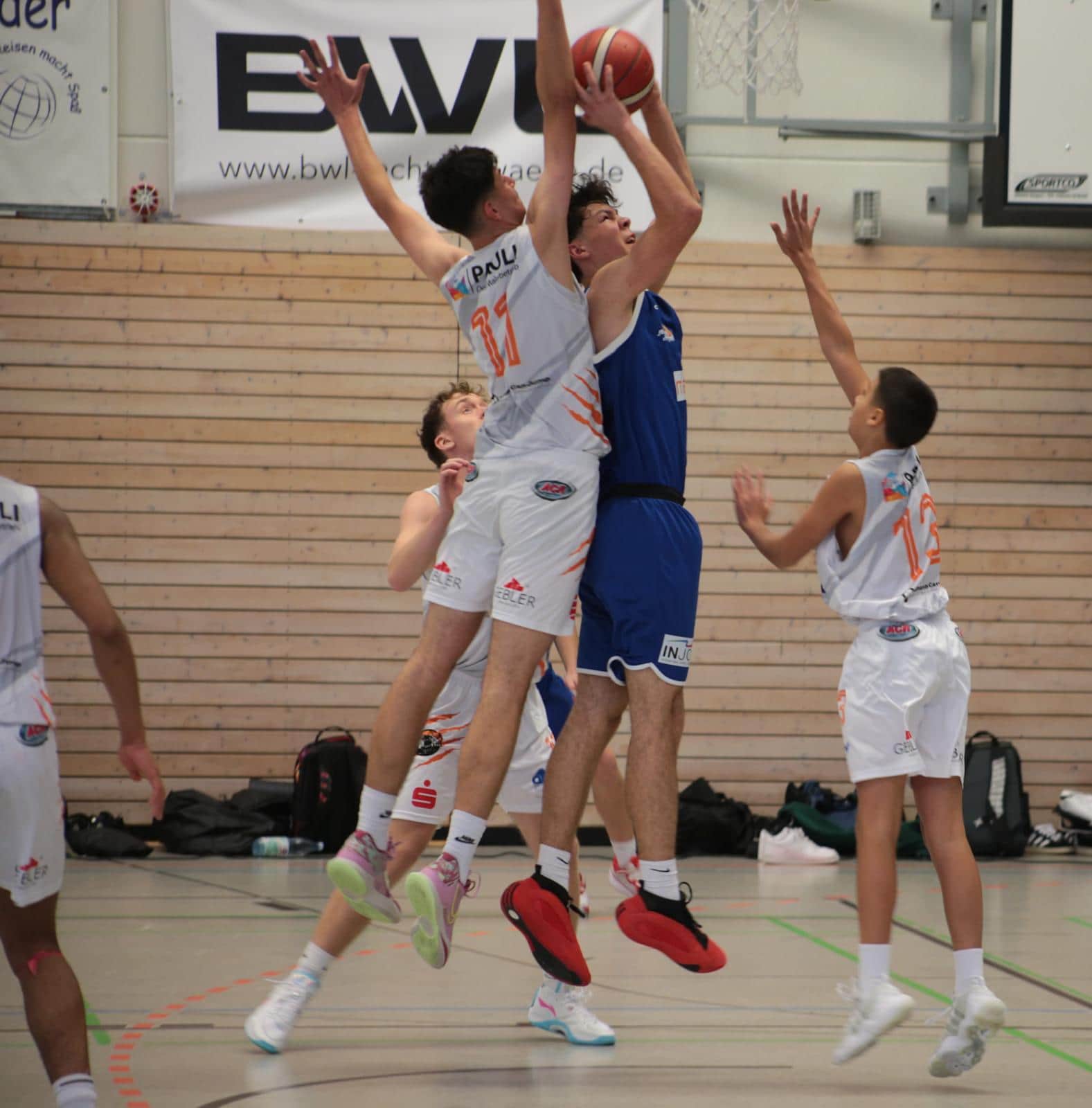 Zwei Basketballspieler im Sprung unter dem Korb. Einer trägt blau, der andere weiß und blockt.