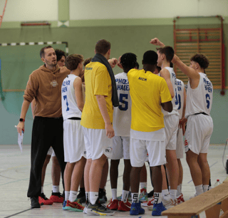 Basketballteam im Kreis, Trainer motiviert. Weiße Trikots, gelbe Shirts, Halle im Hintergrund.