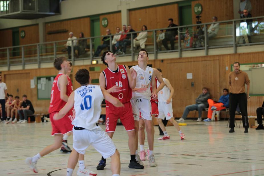 Gruppe junger Männer spielt Basketball in einer Turnhalle. Spannung und Konzentration sind erkennbar.