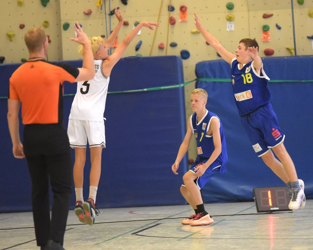 Ein Basketballspieler im weißen Trikot beim Wurf, zwei Verteidiger im blauen Trikot springen.