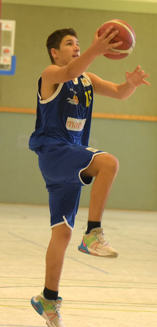 Ein junger Mann im blauen Basketballtrikot springt mit entschlossenem Gesichtsausdruck, hält den Ball.