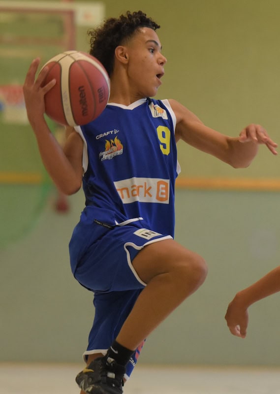 Ein junger Basketballspieler in blauem Trikot springt dynamisch hoch.