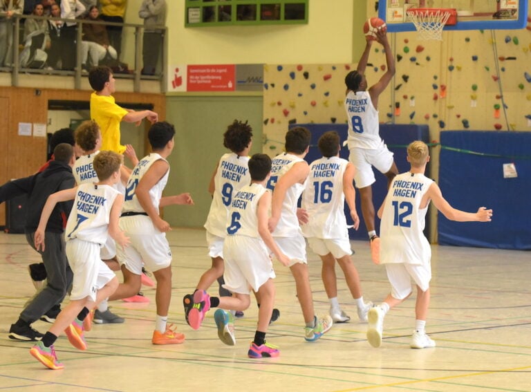 Basketballspiel, Spieler in weißen Trikots, einer springt zum Korb. Zuschauer im Hintergrund.