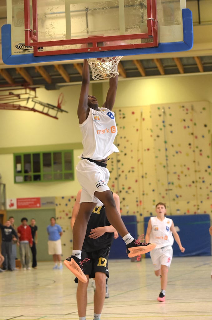 Ein Basketballspieler springt und dunkt kraftvoll. Er trägt ein weißes Trikot und orangefarbene Schuhe.