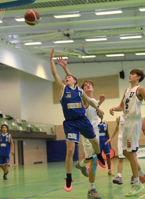 Ein Basketballspieler im blauen Trikot springt und wirft einen Ball, andere Spieler in Aktion.