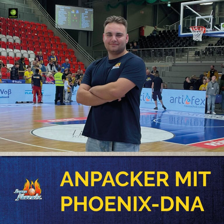 Junger Mann lächelt in Sporthalle mit blauer T-Shirt und Basketballplatz im Hintergrund.