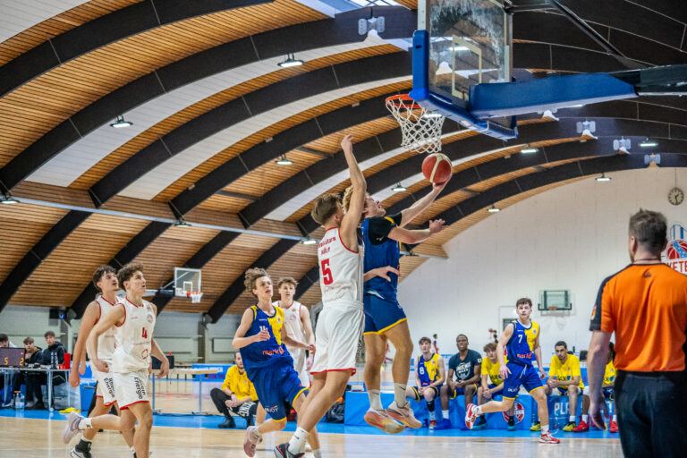 Ein Basketballspiel in einer Sporthalle, ein Spieler in Weiß springt zum Korb, gefolgt von Spielern.