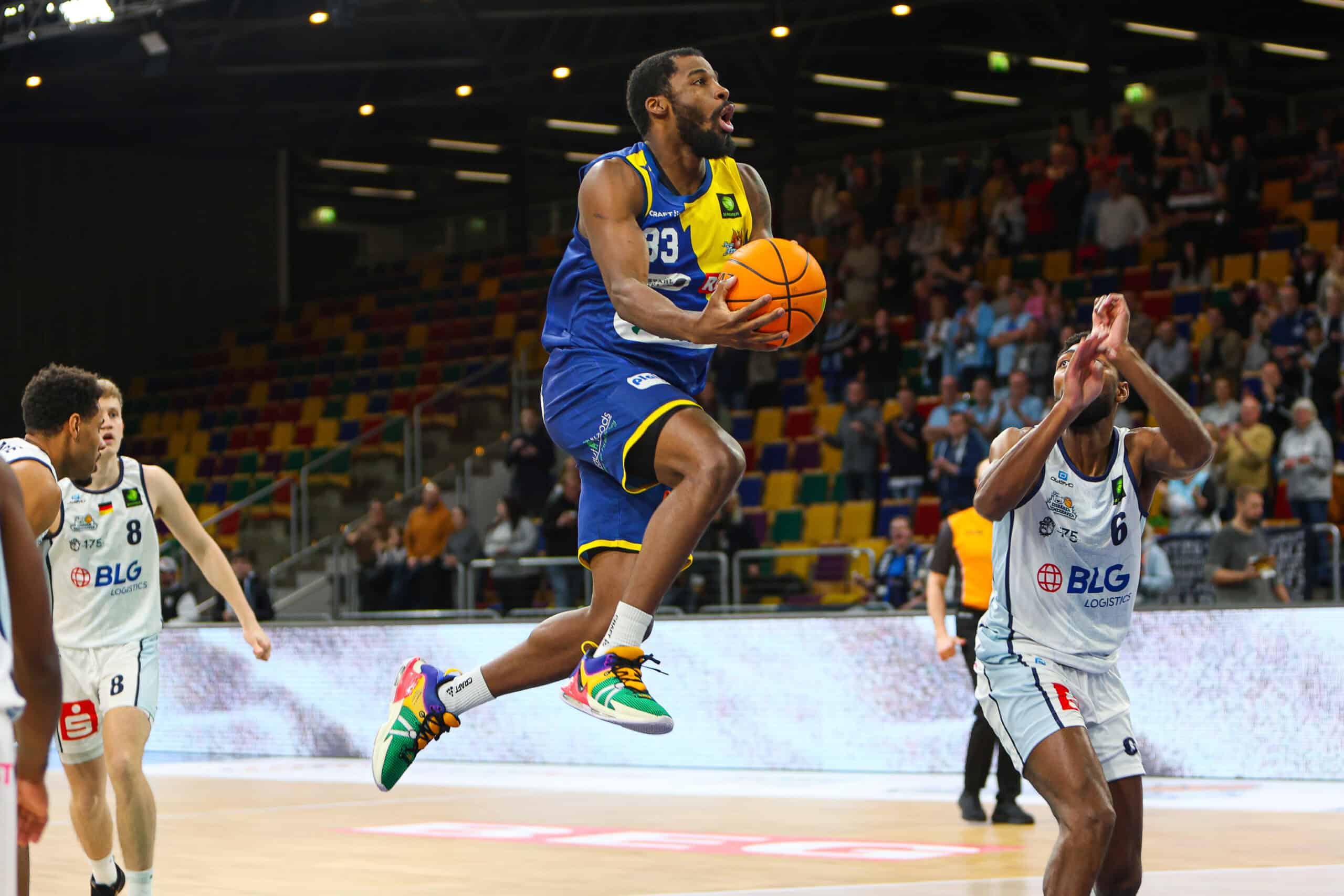 Basketballspieler im blauen Trikot springt mit Ball, andere Spieler in weißen Trikots verteidigen.