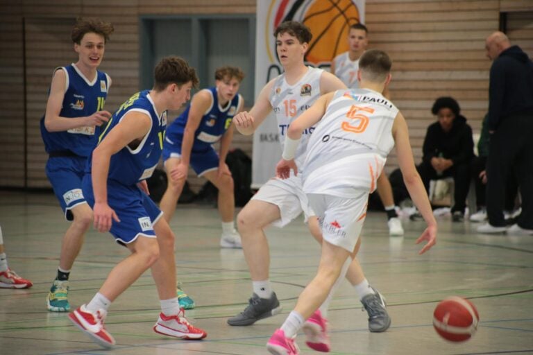 Basketballspiel: Zwei Teams in Blau und Weiß, Spieler dribbeln, angespannte Körperhaltung, Turnhalle.