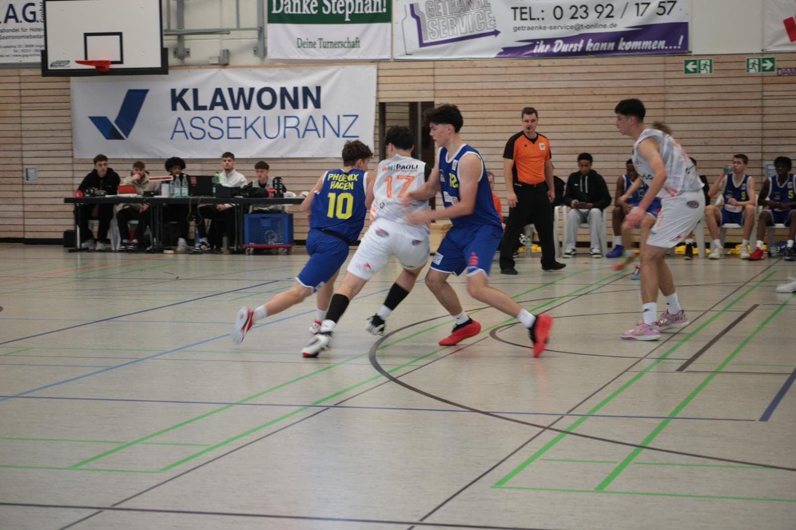 Ein Basketballspiel in einer Turnhalle: Ein Spieler in blauem Trikot wird von zwei Abwehrspielern bedrängt.