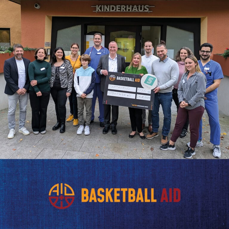 Gruppe lächelnder Menschen vor einem Kinderhaus mit großem symbolischen Scheck, Basketball Aid Logo.