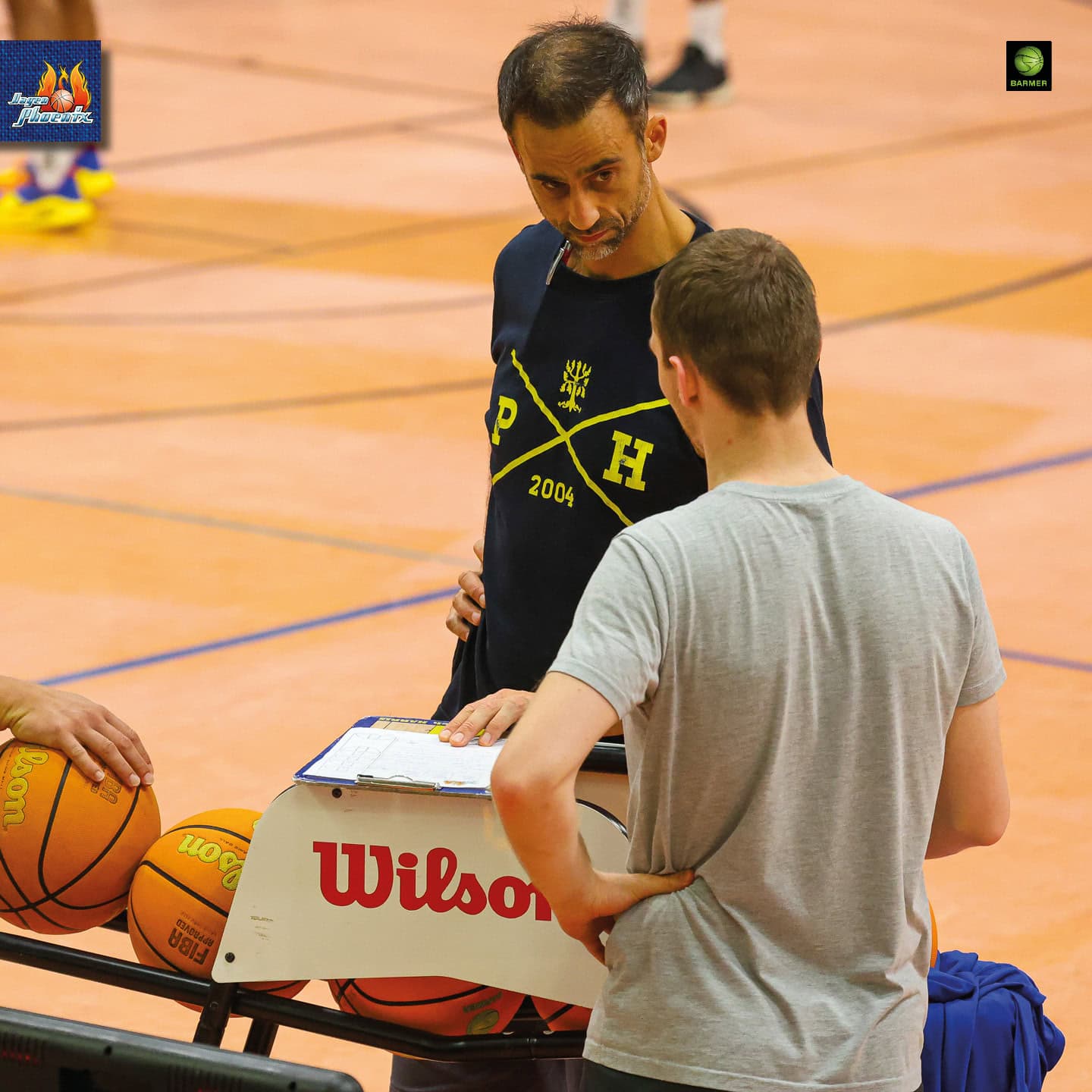 Ein Mann mit nachdenklichem Ausdruck liest ein Clipboard. Basketballkugeln im Hintergrund.