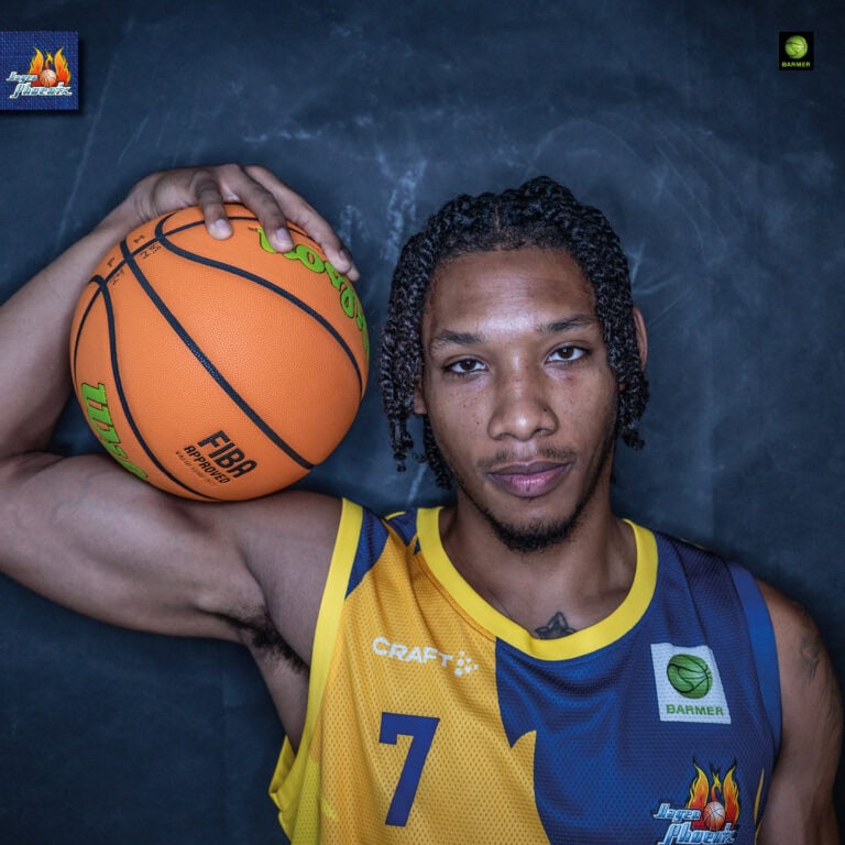 Basketballspieler mit ernstem Blick, trägt gelb-blaues Trikot, hält einen Basketball an der Schulter.