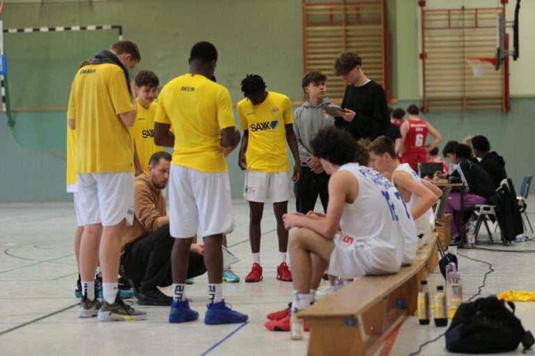 Basketballerstteam in gelben Trikots, besprechen Taktik mit Trainer; Spieler in weißen Trikots sitzen.