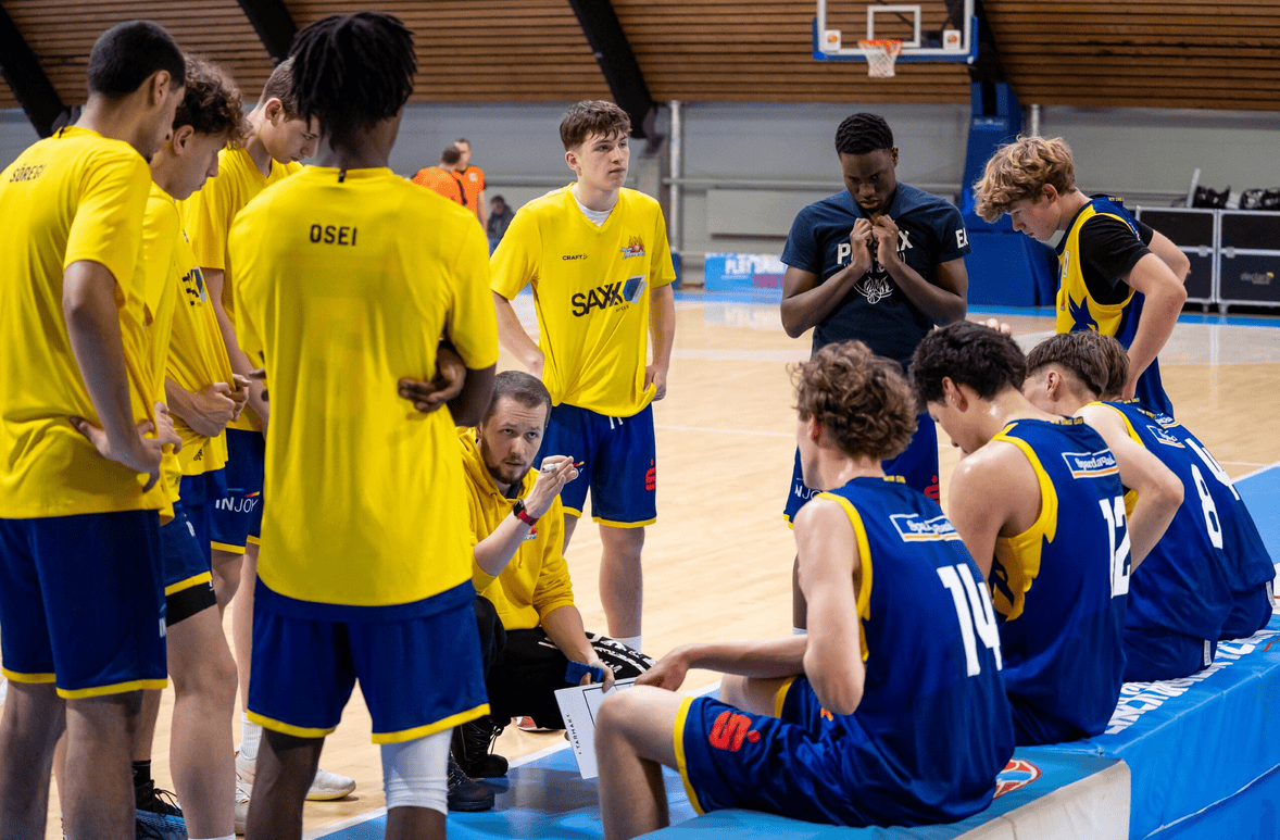 Junger Basketballtrainer diskutiert Taktik mit seinem gelb-blau gekleideten Team auf dem Spielfeld.