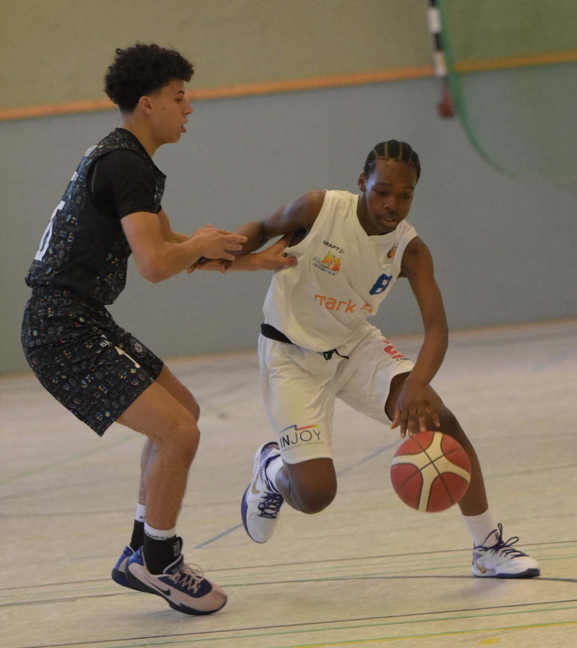 Zwei junge Männer spielen Basketball, einer in weißem Trikot dribbelt, der andere in schwarzem Trikot verteidigt.