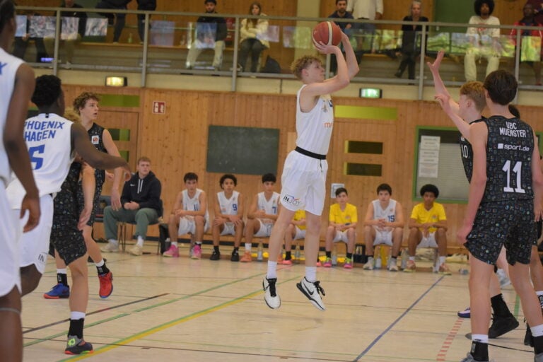 Ein Jugendlicher in weißem Trikot springt und wirft einen Basketball, umgeben von verteidigenden Spielern.