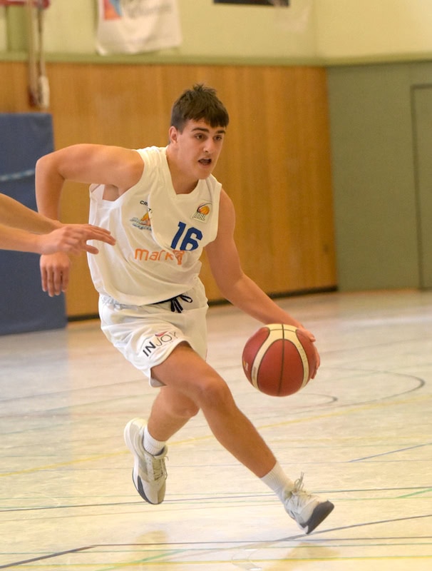 Ein junger Mann spielt Basketball in weißer Sportkleidung und dribbelt konzentriert auf einem Spielfeld.