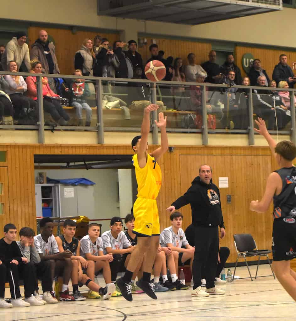 Basketballspieler im gelben Trikot wirft Ball; Zuschauer im Hintergrund; konzentrierter Ausdruck.