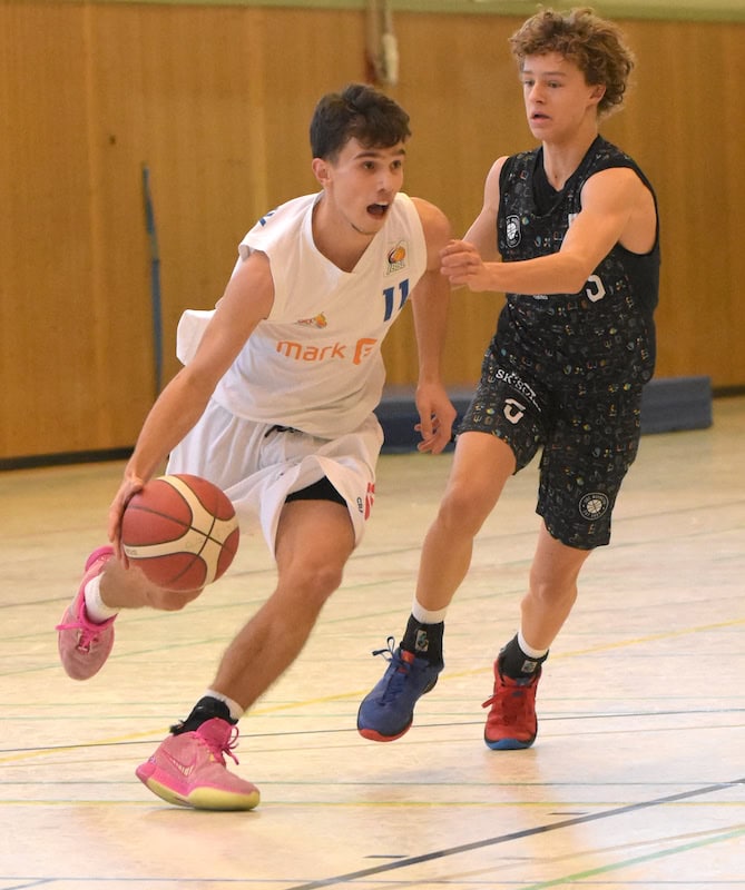 Ein Spieler im weißen Trikot dribbelt intensiv, verfolgt von einem Spieler im schwarzen Trikot.