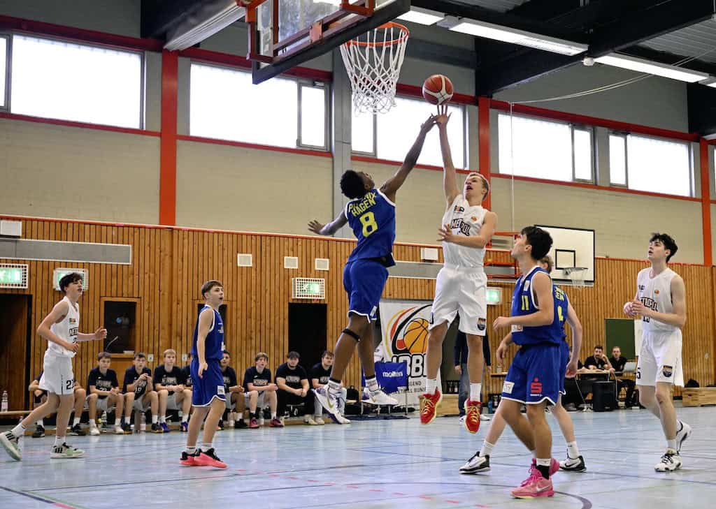 Zwei Basketballspieler springen zum Korb, einer in Blau blockt. Teamkollegen beobachten aufmerksam.