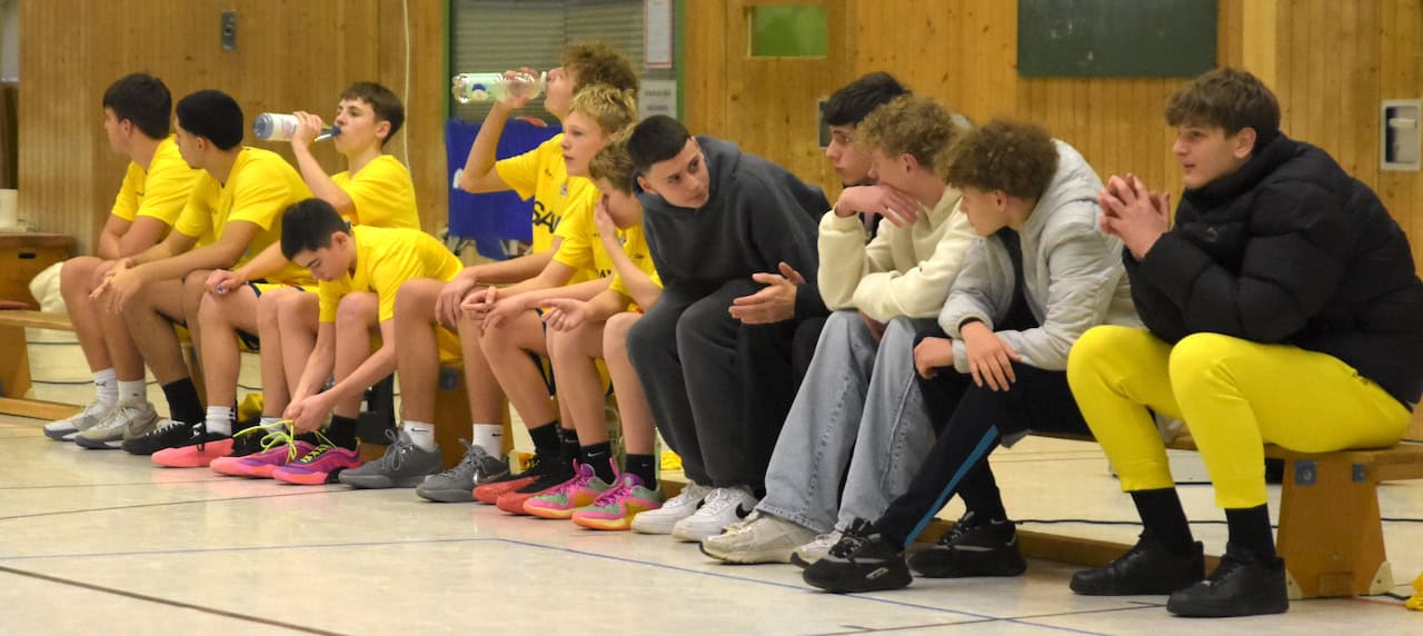 Basketballteam in gelber Kleidung sitzt auf der Bank, einige trinken Wasser, entspannte Stimmung.