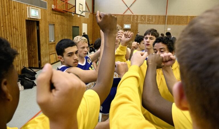 Jugendbasketball-Team im Kreis, entschlossene Mimik, bunte Sportkleidung, in einer Turnhalle.