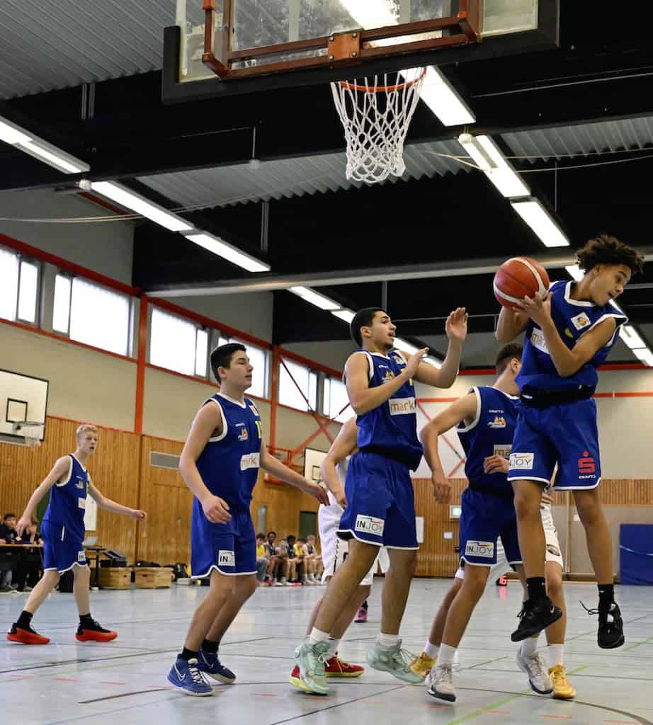 Ein Basketballteam in blauen Trikots spielt in einer Turnhalle, ein Spieler springt nach dem Ball.