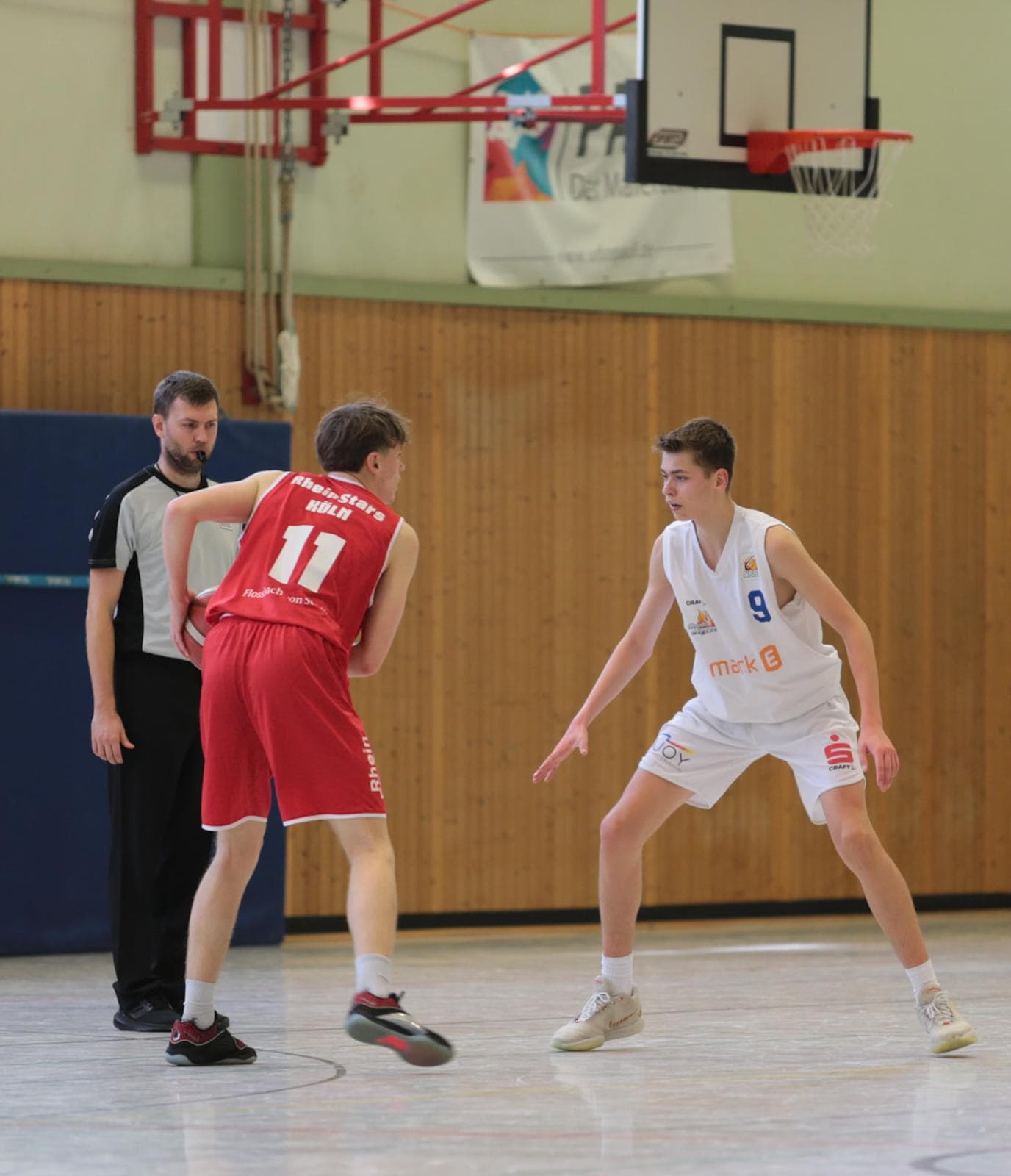 Ein Basketballspiel; Spieler in Rot dribbelt, Spieler in Weiß verteidigt aufmerksam. Schiedsrichter schaut zu.