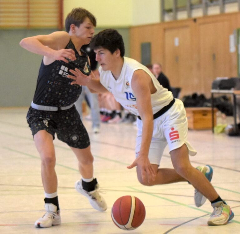 Zwei junge Männer spielen Basketball; einer dribbelt den Ball fokussiert, der andere verteidigt ihn.