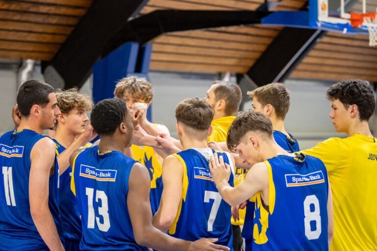 Mehrere Basketballspieler in blauen Trikots beim Team-Meeting in einer Sporthalle.