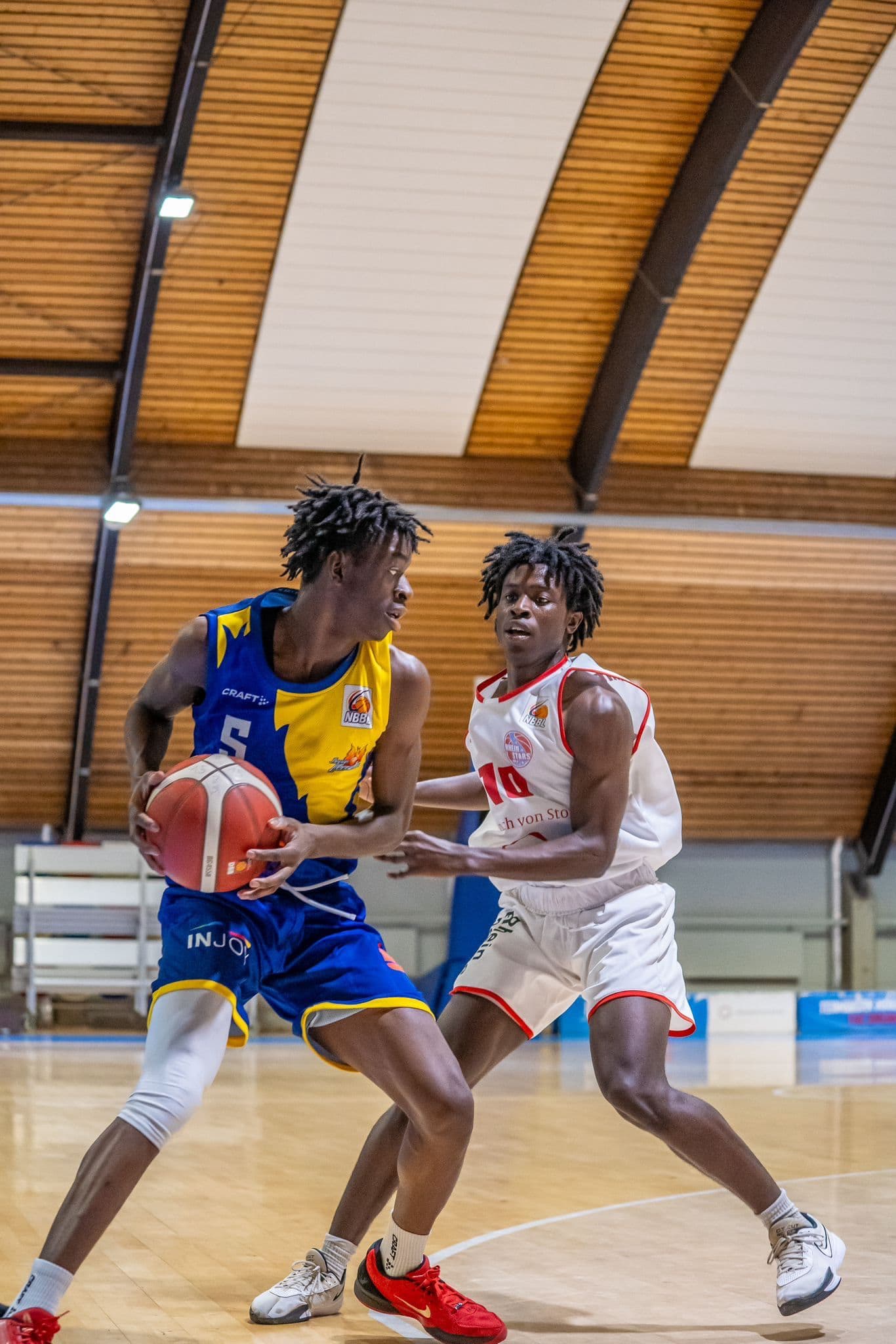 Zwei Basketballspieler im Spiel, einer in blau-gelbem Trikot mit konzentriertem Blick, der andere in weißem Trikot in Verteidigungsposition.