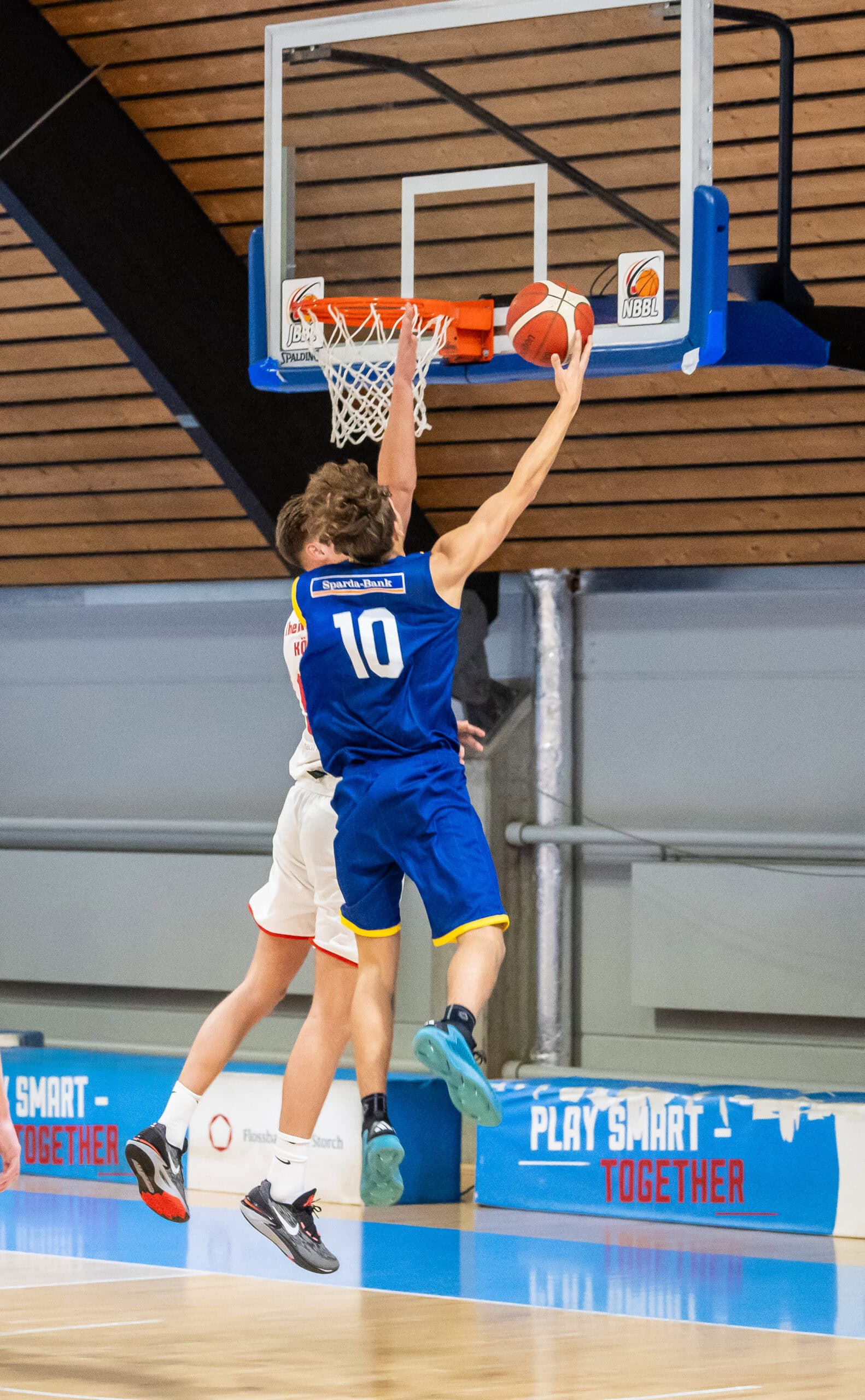 Basketballspieler in blauem Trikot springt zum Korb, Spieler in weiß versucht zu blocken.