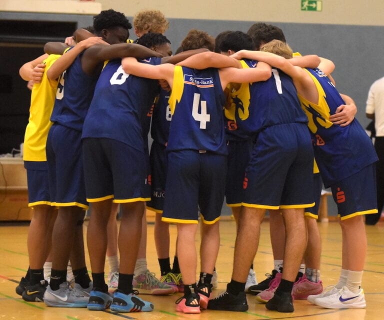 Eine Gruppe von fünf jungen Basketballspielern umarmt sich auf dem Spielfeld. Sie tragen blaue Trikots.