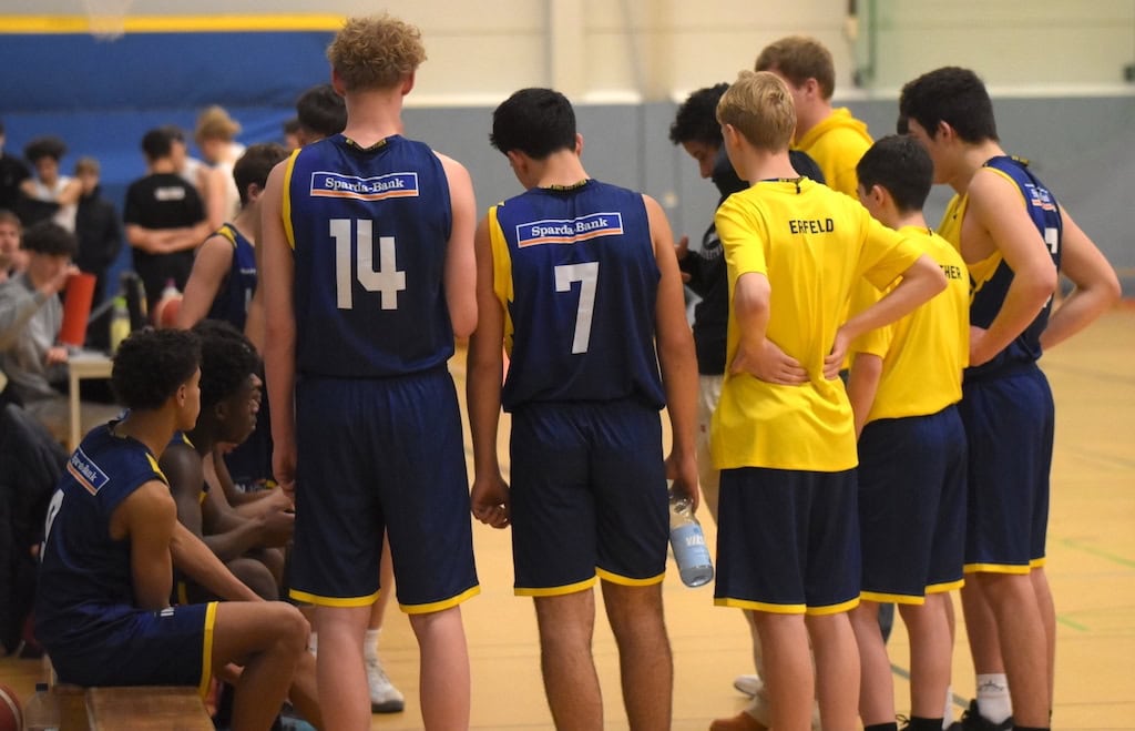 Basketballteam in blauen Trikots mit Nummern, steht im Kreis; Spieler sprechen miteinander.