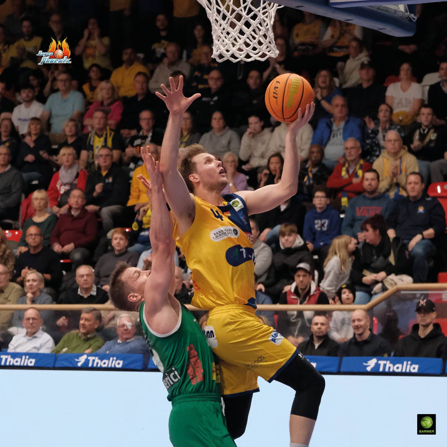 Ein Basketballspieler in gelbem Trikot macht einen Korbleger, während ein Verteidiger in grün zuschaut.