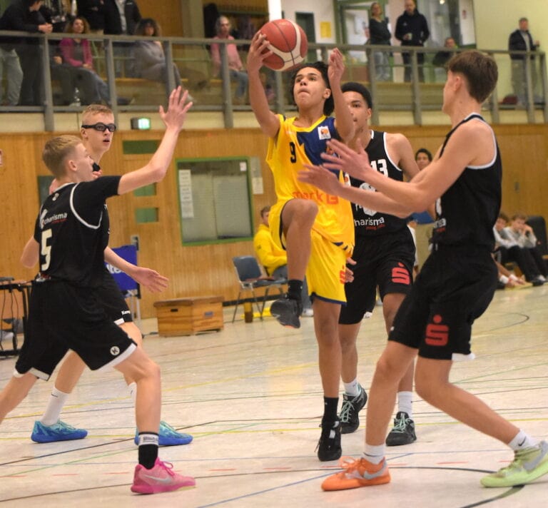 Ein junger Mann in gelbem Trikot beim Basketballwurf, umgeben von Gegenspielern in schwarzen Trikots.