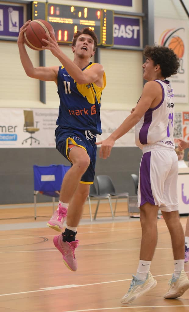 Ein Basketballspieler im blauen Trikot springt zum Korb; ein Gegner im weißen Trikot steht daneben.