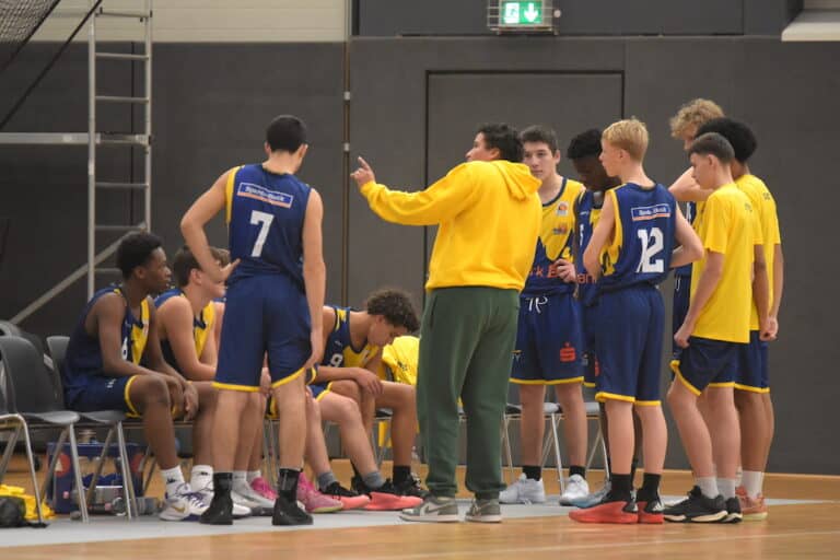 Ein Trainer spricht ernst mit einem Basketballteam in blauen und gelben Trikots in der Halle.