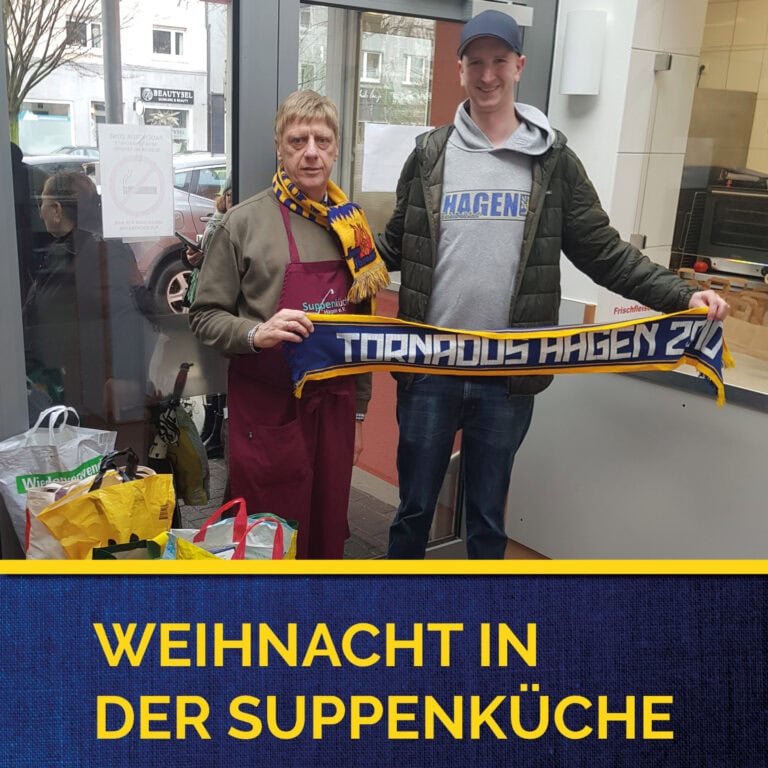 Zwei Männer lächeln, einer mit Schürze, halten ein blau-gelbes Banner in einer Suppenküche.