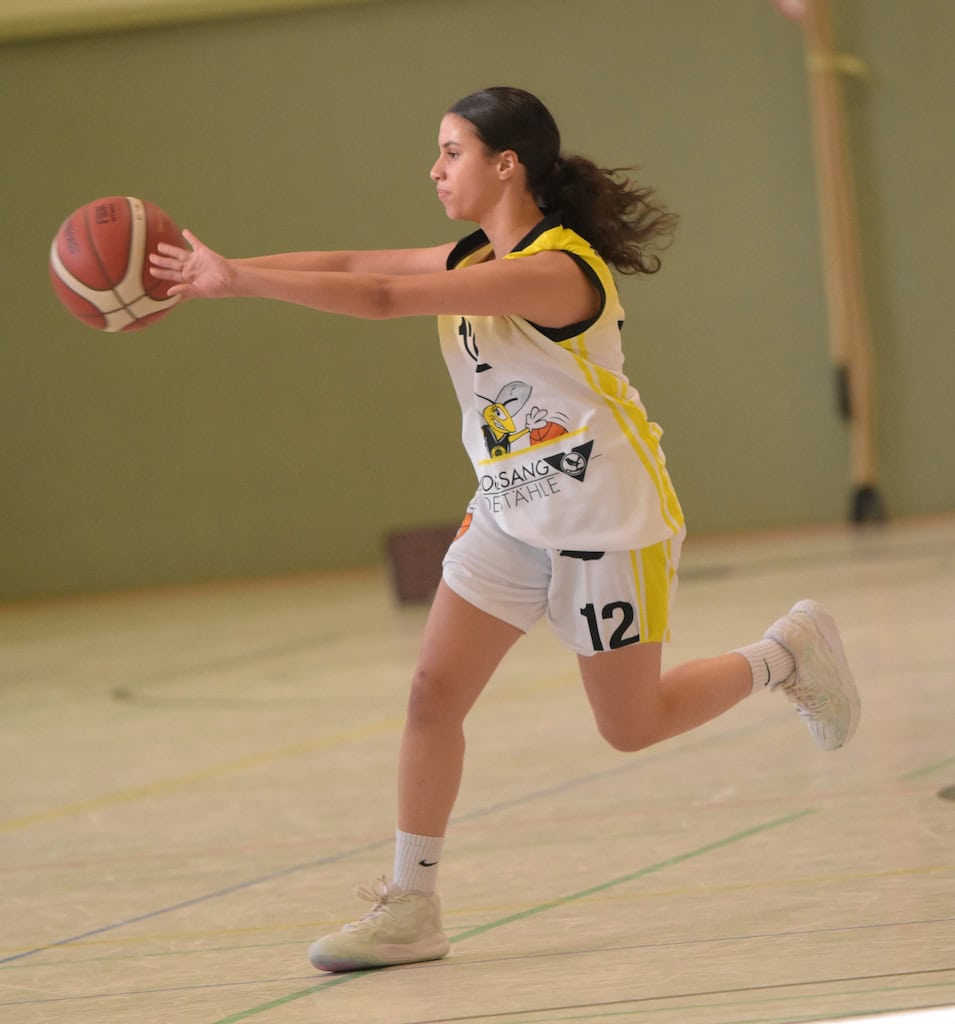 Eine junge Basketballspielerin in Aktion, konzentriert, trägt ein weiß-gelbes Trikot mit der Nummer 12.