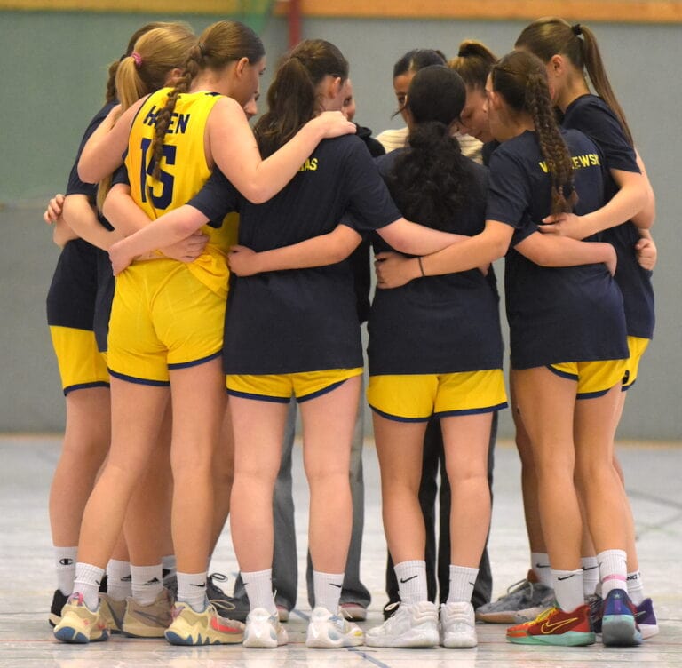 Eine Gruppe von Handballspielerinnen in gelb-blauer Teamkleidung umarmt sich auf dem Spielfeld.