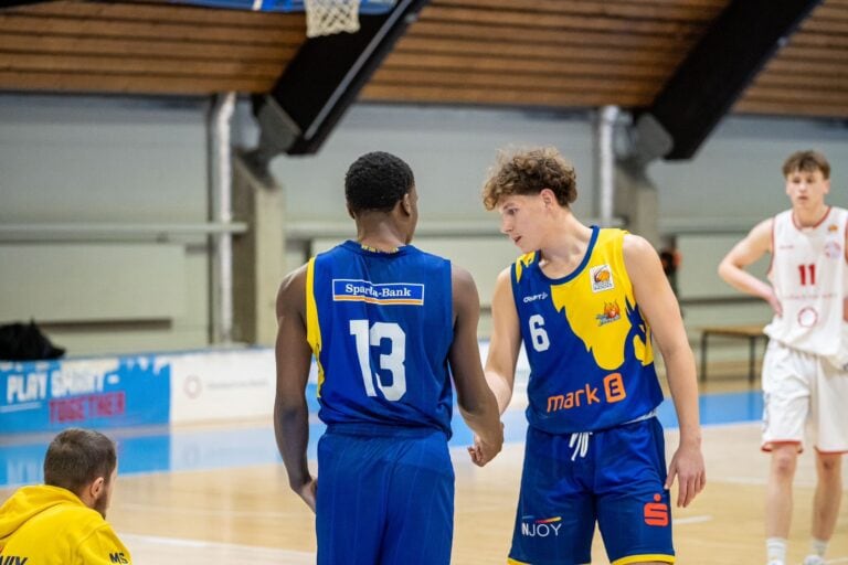 Zwei Basketballspieler im blauen Trikot tauschen während eines Spiels einen Handshake aus.